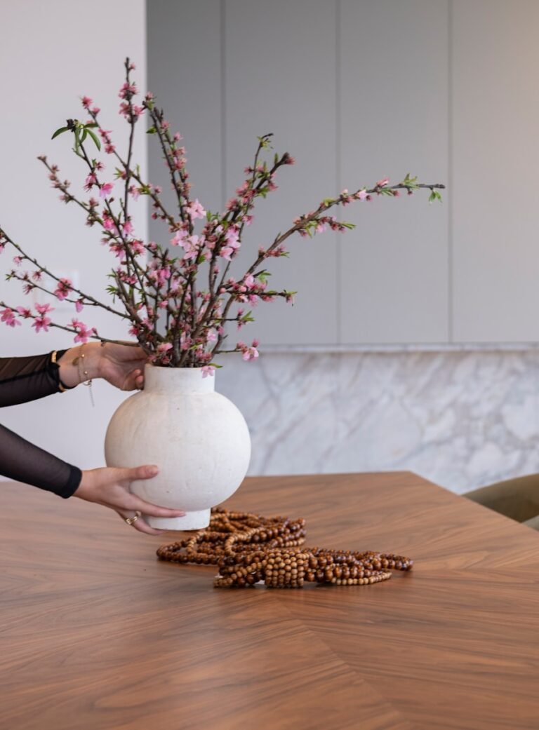 Hands arranging blooming branches in a white vase on table.
